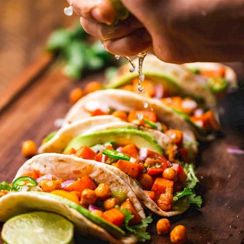 The image shows a hand squeezing lime juice over a row of tacos filled with chickpeas, avocado, and vegetables, garnished with cilantro and served on a wooden board.