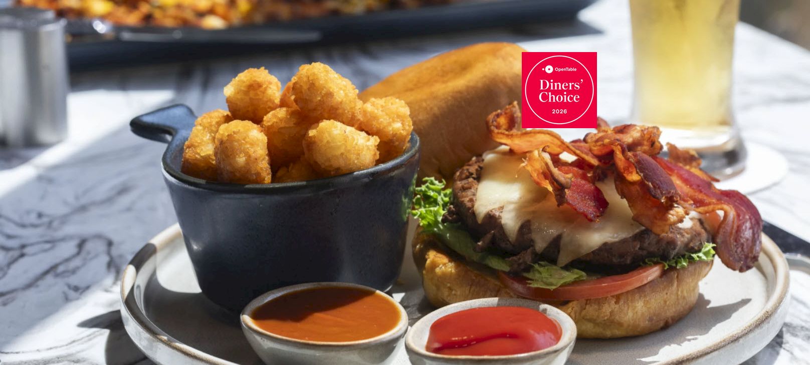 A bacon cheeseburger with crispy fried bites and two dipping sauces on a plate.