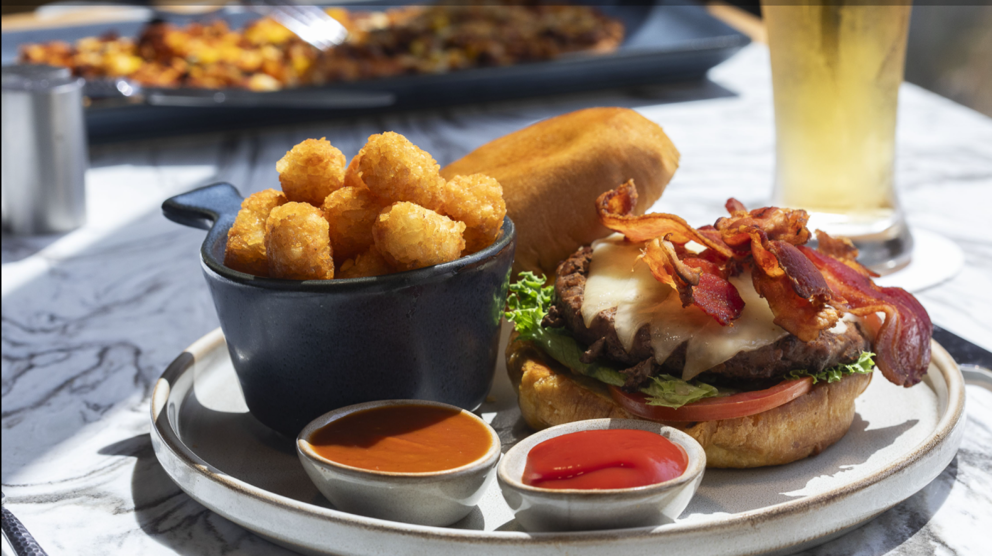 The image shows a plate with a bacon cheeseburger, a side of tater tots, and two dipping sauces. A beverage and another dish are also visible in the background.