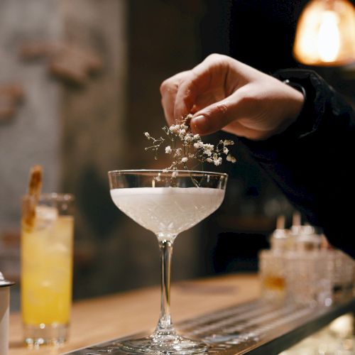 A person is garnishing a cocktail in a coupe glass with a sprig of herbs at a bar. Another cocktail is in the background.