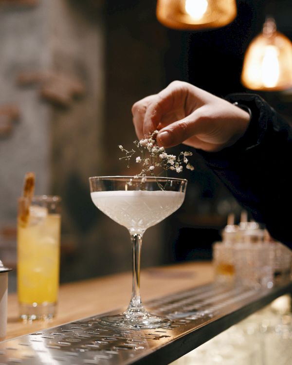 A person is garnishing a cocktail in a coupe glass with a sprig of herbs at a bar. Another cocktail is in the background.