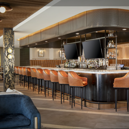 This image shows a modern bar area with orange bar stools, two large TVs above the bar, and a stylish interior design with wood and gold accents.