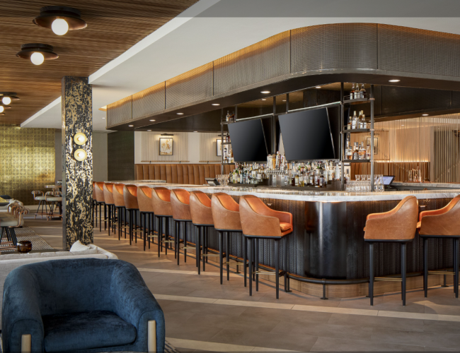 This image shows a modern bar area with orange bar stools, two large TVs above the bar, and a stylish interior design with wood and gold accents.