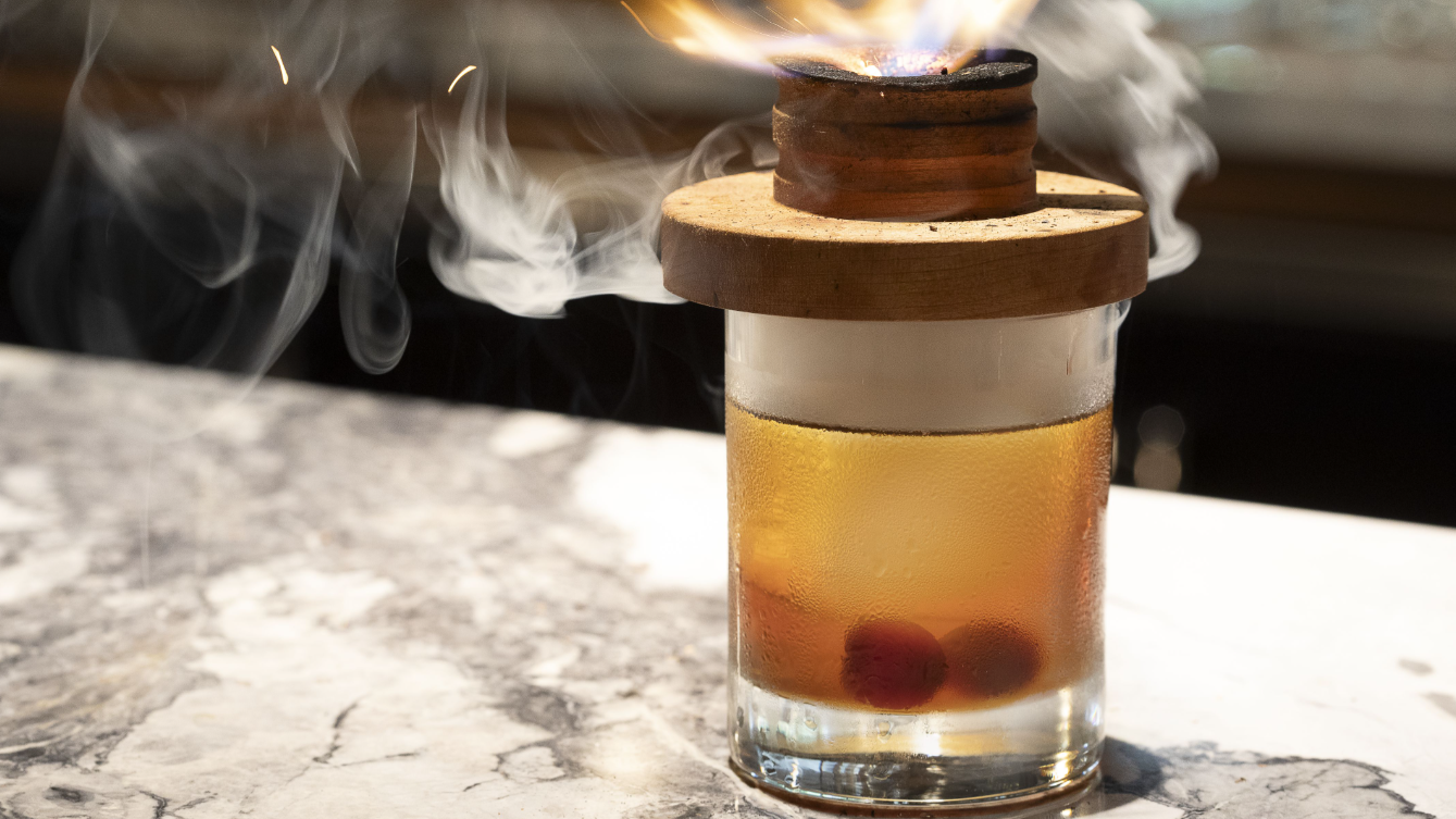 A cocktail glass with cherries inside, a smoky and fiery wooden cap on top, sitting on a marble surface.