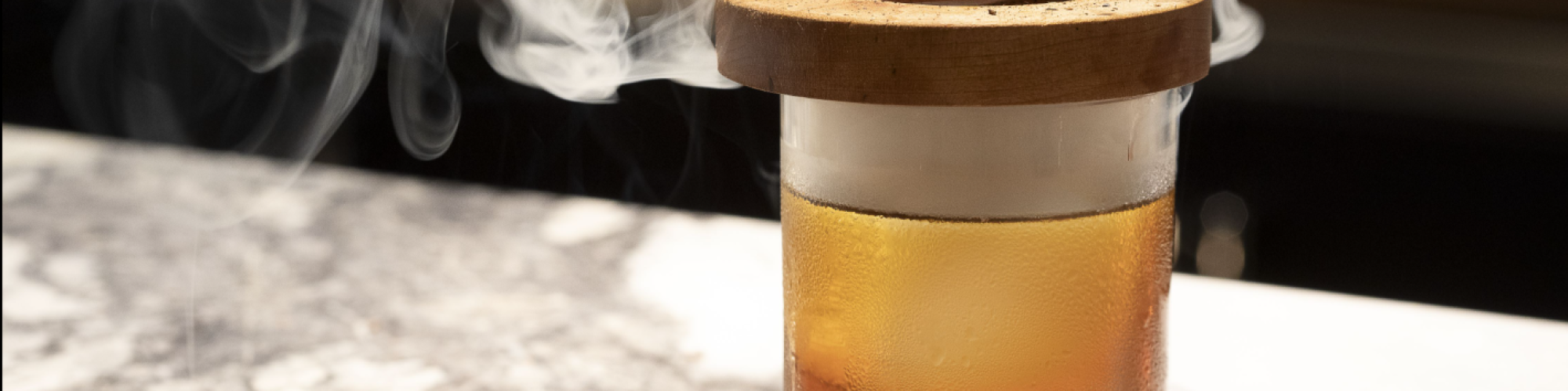 A cocktail glass with cherries inside, a smoky and fiery wooden cap on top, sitting on a marble surface.