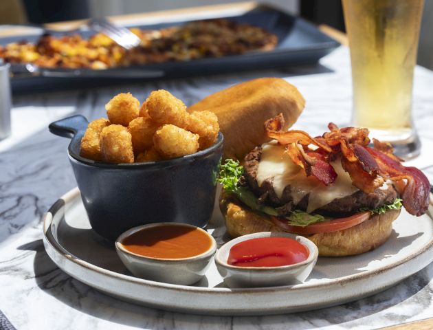 A burger with bacon and cheese, tater tots, two dipping sauces, and a tall glass of beer on a marble table at a restaurant.