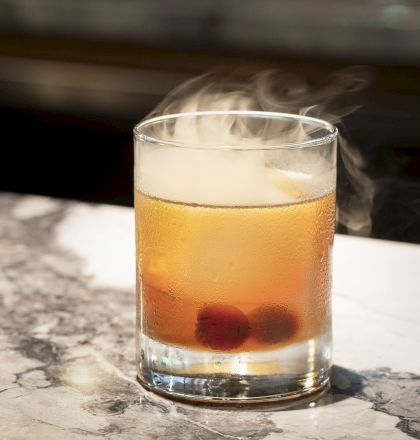 A cocktail in a glass with two cherries at the bottom, sitting on a marble surface, with steam or smoke rising from the drink, indicating it is cold.