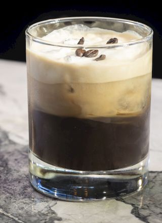 A glass of layered iced coffee topped with foam and coffee beans sits on a marbled surface, creating a visually appealing drink and background.