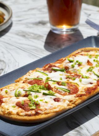 The image shows a rectangular flatbread pizza topped with cheese and green garnishes on a plate, set on a marble table with a drink and utensils.