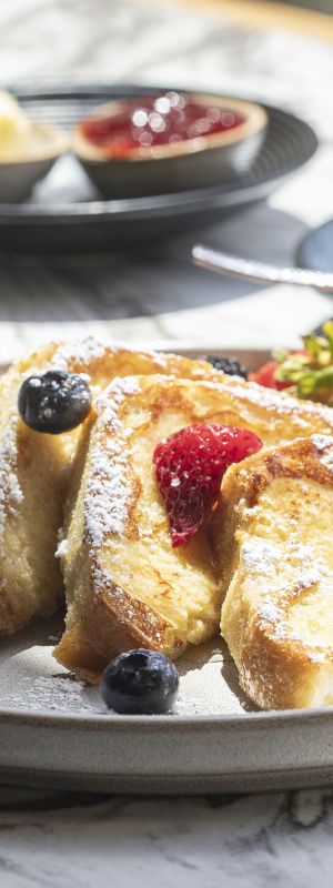 A plate of French toast with berries and powdered sugar, accompanied by a cup of coffee, butter, and jam in the background.