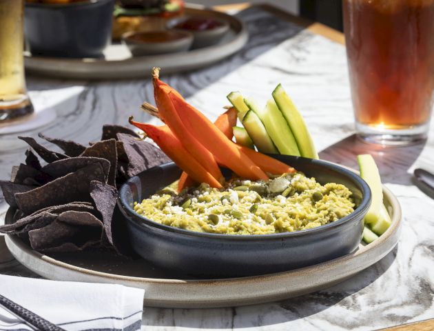 A plate with blue corn chips, guacamole, carrot, and cucumber sticks, next to two drinks on a marble table.