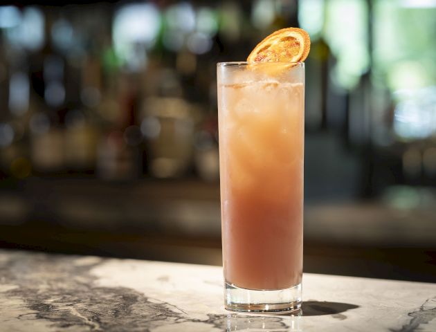 A tall glass with an iced pink cocktail garnished with a dehydrated orange slice; the drink is placed on a marble surface with a blurred background.