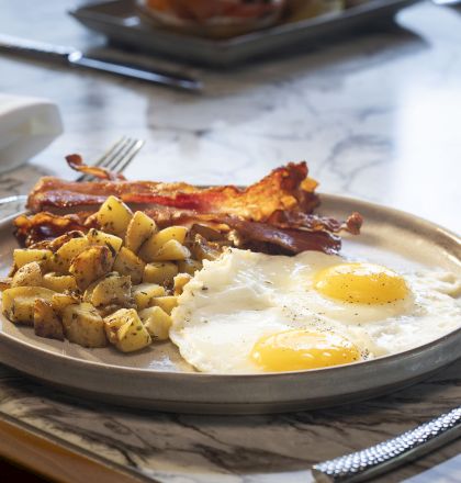 A plate with two sunny-side-up eggs, bacon, and diced potatoes served on a marble table with a glass of orange juice and silverware.