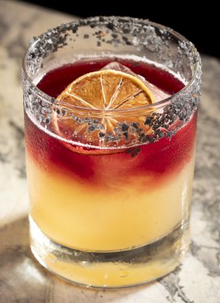 A glass filled with a layered cocktail, featuring a slice of dried citrus and a salted rim, placed on a marble surface.