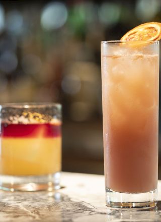 The image shows two cocktails on a bar, one short drink with a reddish layer and one tall iced drink garnished with an orange slice.