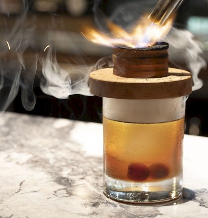 The image shows a cocktail glass with a wooden lid, a smoking flame on top, and cherries at the bottom of the drink, placed on a marble surface.