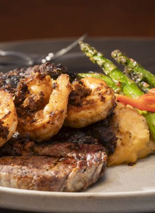 A plate with grilled steak topped with shrimp, served with mashed potatoes, asparagus, and red peppers, with a drink in the background.