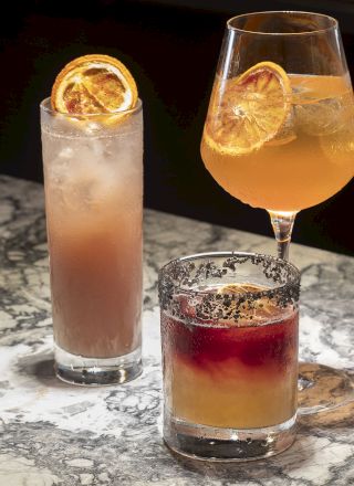 Three cocktails on a marble counter, each garnished with a dried citrus slice, in different glasses, highlighting their colors and presentation.