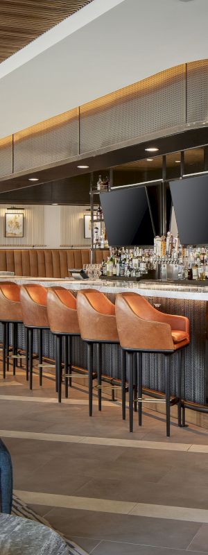 A modern bar with orange barstools, two large TV screens, stylish lighting, and seating areas featuring blue and gray chairs and tables finishes the scene.