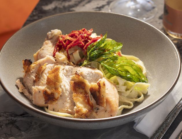 A pasta dish topped with grilled chicken slices, basil leaves, and what appears to be diced tomatoes or red pepper with a drink and cutlery beside it.