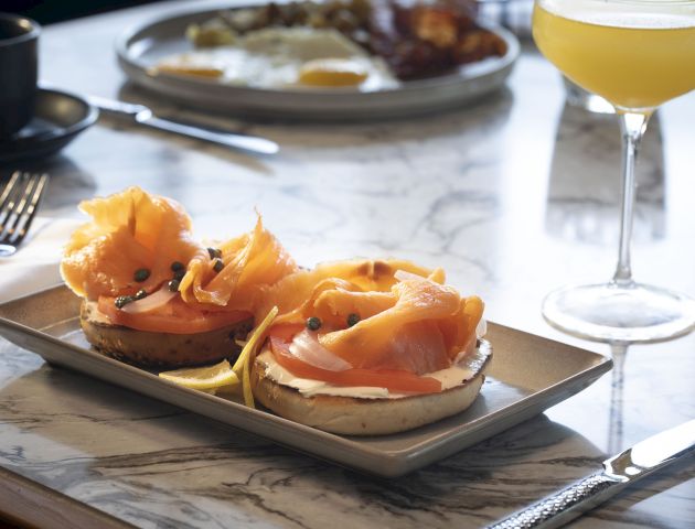 The image shows a smoked salmon bagel with cream cheese, capers, and sliced tomato on a plate, accompanied by a glass of orange juice and a coffee cup.