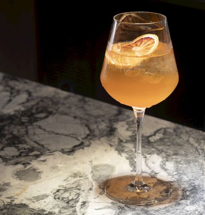 A glass with a light orange-colored cocktail garnished with a citrus slice rests on a marbled surface.