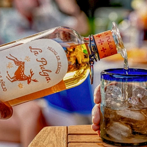 A person pours tequila from a bottle labeled "La Pulga" into a glass with ice, set on a wooden surface in an outdoor setting.