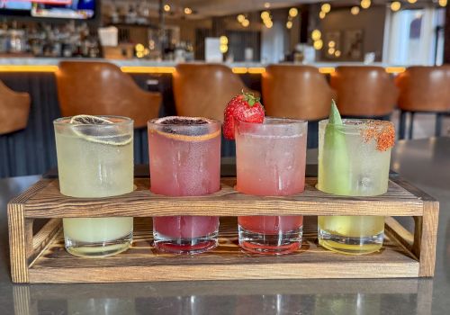 Four colorful frozen cocktails in tall glasses sit on a wooden tray in a cozy bar, with warm string lights behind.