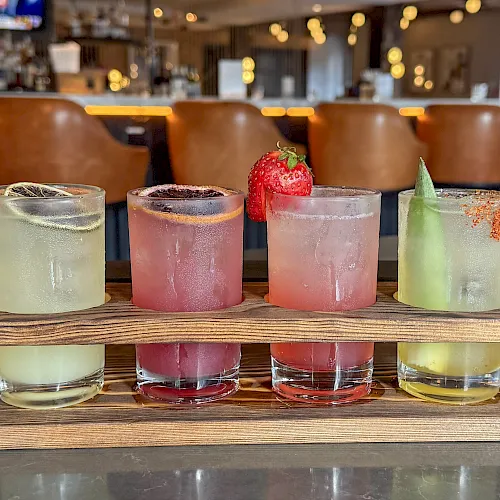 Four colorful frozen cocktails in tall glasses sit on a wooden tray in a cozy bar, with warm string lights behind.