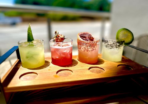 A wooden flight of four colorful cocktails on a tray: lime green, red, pink with chili salt rim, and pale yellow with a cucumber slice, sunny outdoors.
