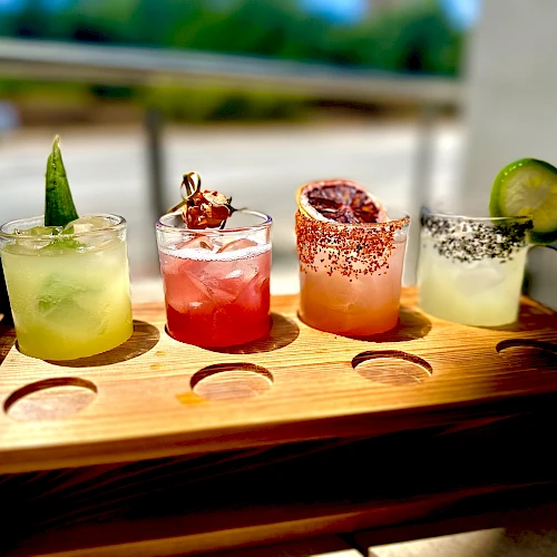 A wooden flight of four colorful cocktails on a tray: lime green, red, pink with chili salt rim, and pale yellow with a cucumber slice, sunny outdoors.